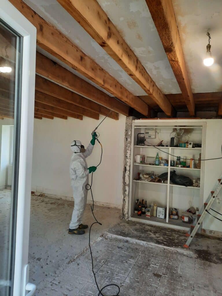 Technicien CBH en tenue de protection appliquant un traitement de charpente par pulvérisation sur des solives, près de Rennes